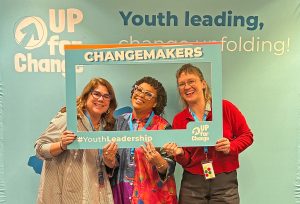 Three individuals, Courtney Welsh (Chief Executive Officer), Kat McIntosh (Content Creation Lab Program Coordinator), and Katie Cox (Senior Program Manager), stand behind a large blue and orange "Changemakers" photo frame with "#YouthLeadership" written at the bottom at an Up For Change event, smiling at the camera.