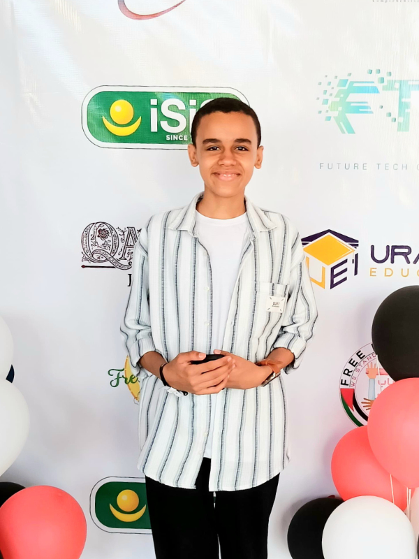 A teenage boy with medium brown skin and short black hair stands smiling in front of an event backdrop with various logos and colorful balloons. He is wearing a striped collared shirt, a white t-shirt, and black pants.