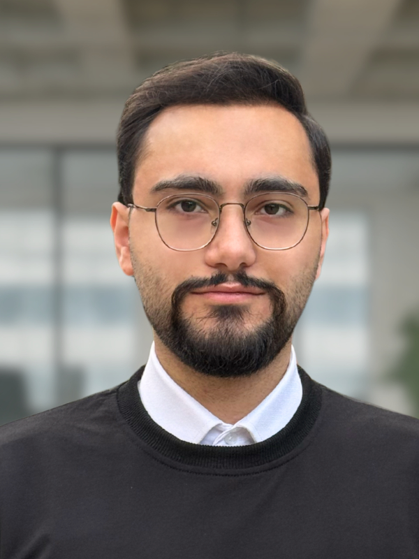 A portrait of a young man wearing glasses, with a light beard and a short hairstyle. He stands in front of a blurred office background