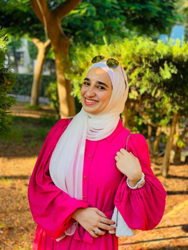 A young woman with fair skin smiles broadly at the camera against a background of green trees. She is wearing a hot pink dress, a white hijab, a watch, a purse, two silver rings, and sunglasses on her head.
