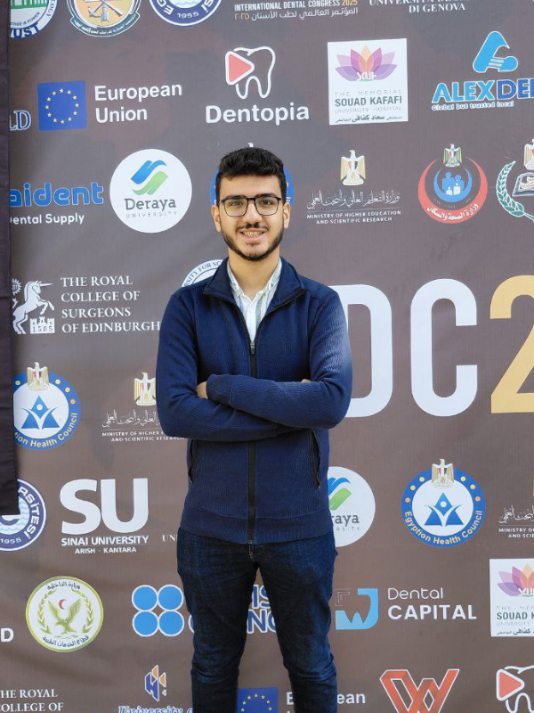 A smiling young man with light brown skin, short black hair, and a black beard stands with his arms crossed in front of an event backdrop. He is wearing blue jeans, a blue sweater, a white shirt, white shoes, and black eyeglasses.