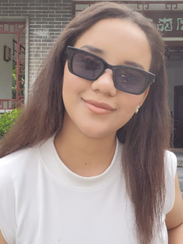 A young woman with long brown hair looks faces the camera with a slight smile, wearing black sunglasses with rectangular frames, a white shirt, and a small gold earring.