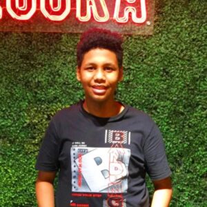 A young man with short, dark curly hair smiling in front of a green foliage wall with a red neon sign partially visible at the top. He is wearing a black graphic t-shirt.