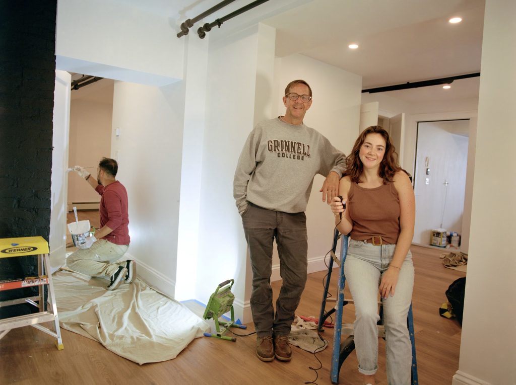 Young girl with pale skin and brown hair standing next to man with light olive skin and glasses in the middle of a living room being renovated