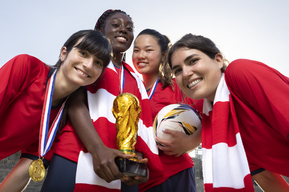 A low-angle close-up of four diverse, smiling female soccer players posing closely together after a victory. They wear red jerseys, gold winner's medals, and red-and-white striped scarves. Two players in the center hold a gold trophy resembling the World Cup, while another player holds a soccer ball.